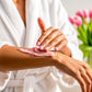 Person applying B.o.T.B. Body cherry body butter to their arm with a blurred background of flowers
